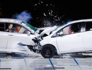 Two white cars in a car testing facility smashing together in a head-on collision as the crash test dummies hit the inflated air bags.